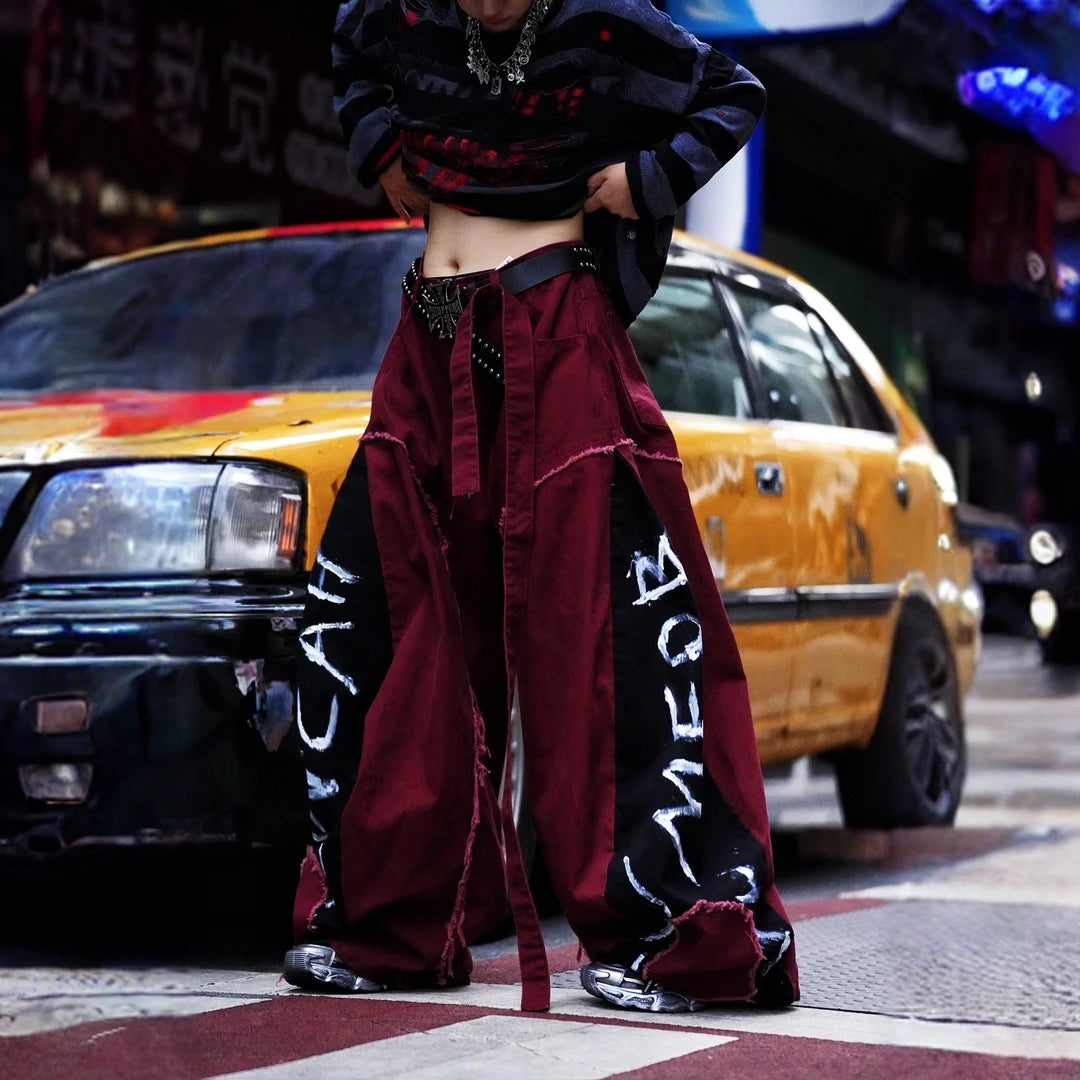 Sanhoostudio denim pants featuring red-toned detailing, heavyweight washed construction, distressed effects, patchwork elements, and a relaxed wide leg scythe silhouette. Designed for statement streetwear styling with rugged texture, comfortable movement, and bold everyday appeal.