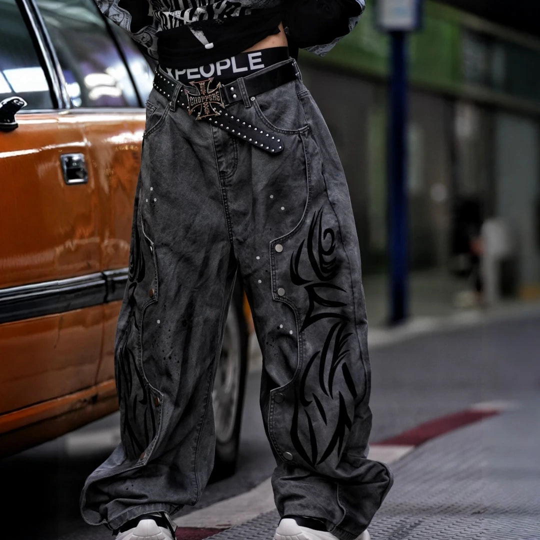 Sanhoostudio denim pants featuring grey and black totem-inspired detailing, heavyweight washed construction, distressed effects, and patchwork elements with a wide leg scythe silhouette. Designed for bold streetwear styling with relaxed comfort and distinctive urban character.
