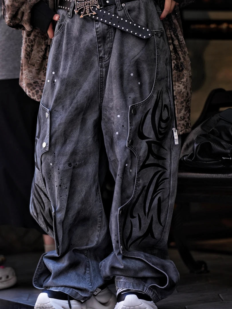 Sanhoostudio denim pants featuring grey and black totem-inspired detailing, heavyweight washed construction, distressed effects, and patchwork elements with a wide leg scythe silhouette. Designed for bold streetwear styling with relaxed comfort and distinctive urban character.