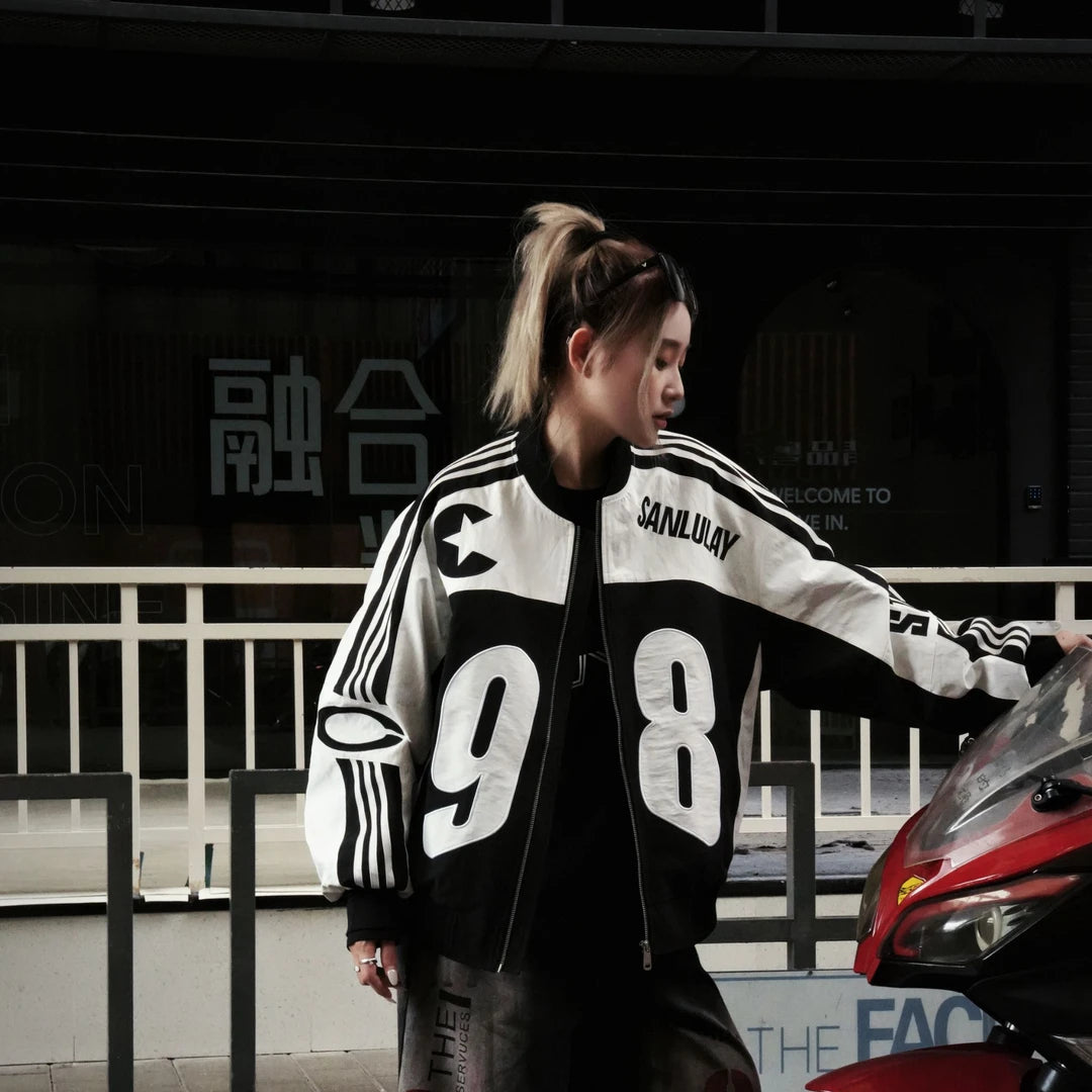 Sanhoostudio bomber jacket featuring black and white colorblock panels, number and letter graphics, a zip-up front, and a cropped racing-inspired silhouette. Designed for early fall streetwear with sporty motorcycle character and versatile everyday layering.