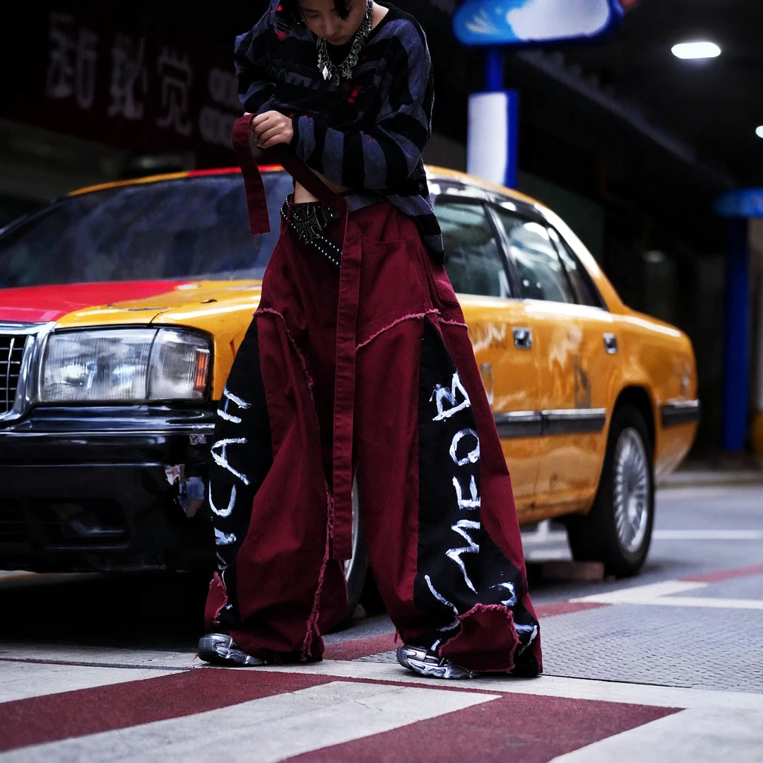 Sanhoostudio denim pants featuring red-toned detailing, heavyweight washed construction, distressed effects, patchwork elements, and a relaxed wide leg scythe silhouette. Designed for statement streetwear styling with rugged texture, comfortable movement, and bold everyday appeal.