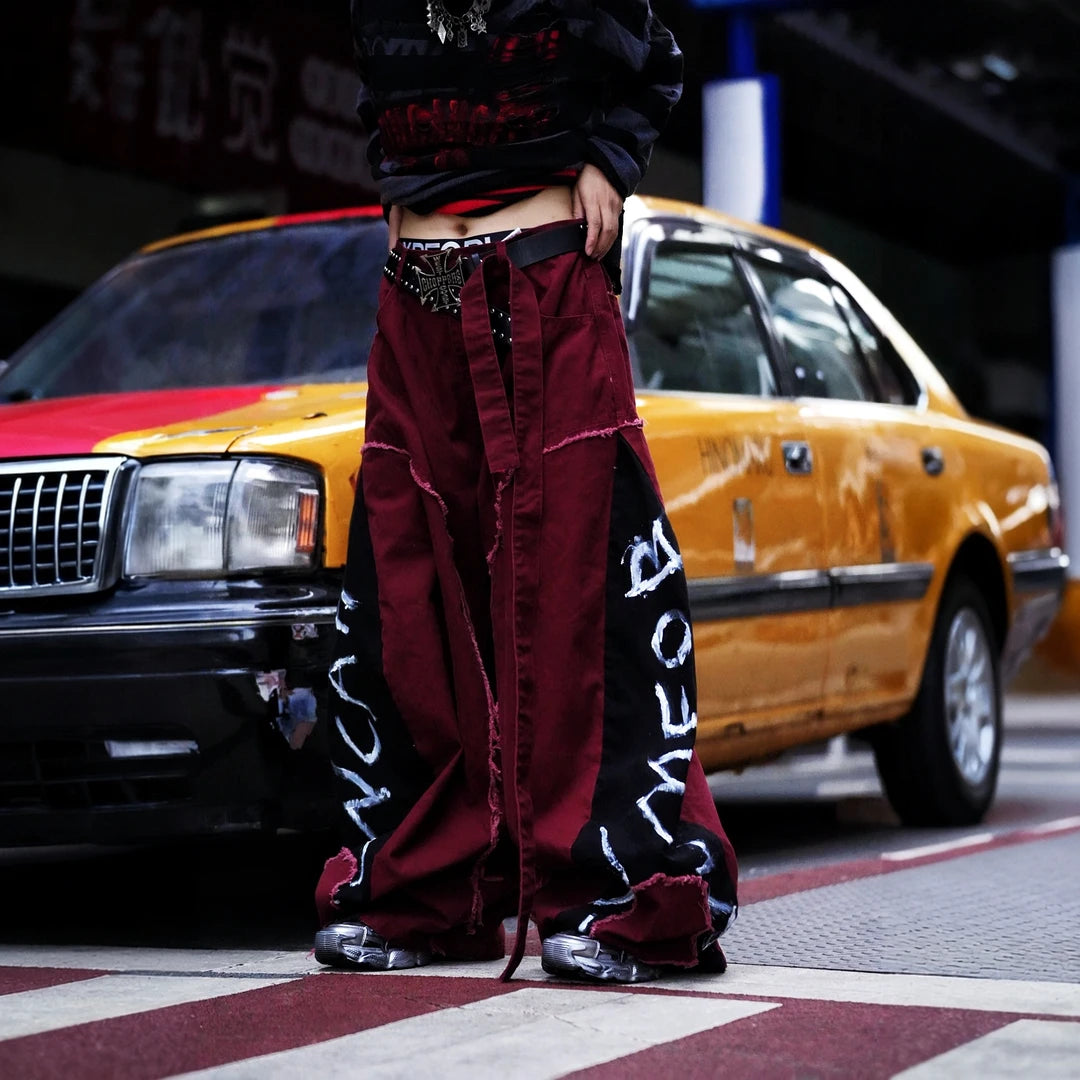 Sanhoostudio denim pants featuring red-toned detailing, heavyweight washed construction, distressed effects, patchwork elements, and a relaxed wide leg scythe silhouette. Designed for statement streetwear styling with rugged texture, comfortable movement, and bold everyday appeal.