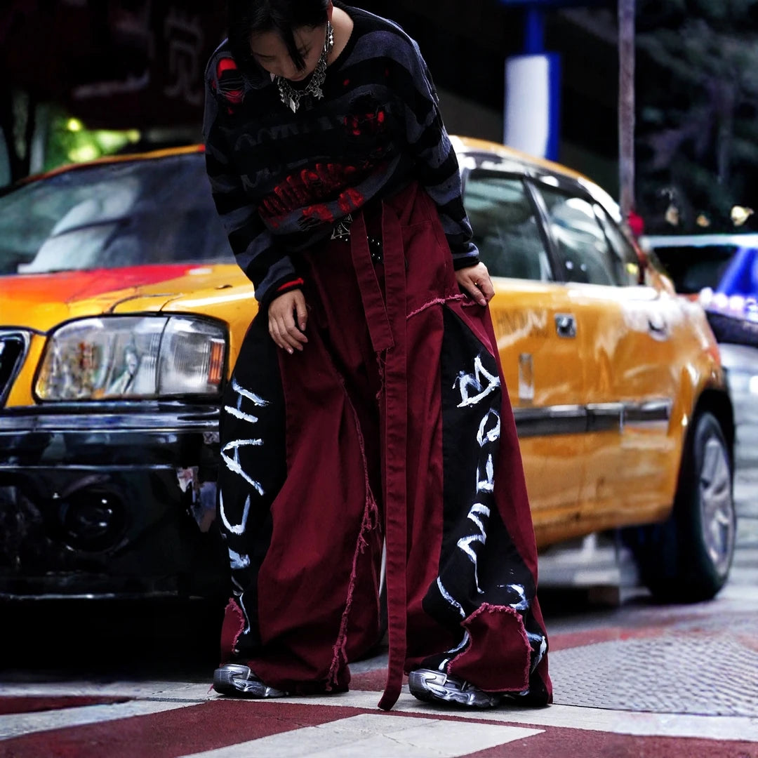 Sanhoostudio denim pants featuring red-toned detailing, heavyweight washed construction, distressed effects, patchwork elements, and a relaxed wide leg scythe silhouette. Designed for statement streetwear styling with rugged texture, comfortable movement, and bold everyday appeal.